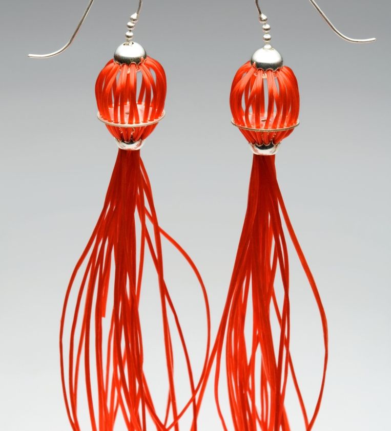 Medusa Ohrringe. Material: Silber, Gummischnüre rot. Der Körper dieser leichten Ohrringe wird durch viele rundlich angeordnete Gummischnüre geformt, die in einem tentakelartigen Schweif ausläuft.
