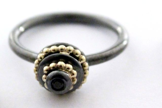 Ring aus geschwärztem Silber, hat einen törtchenförmigen Oberteil. Auf diesem ist auf zwei Ebenen ein Kugeldraht aus 14 karätigem Gold aufgelötet. Oben in der Mitte bildet ein facettierter, schwarzer Spinell den Abschluss.