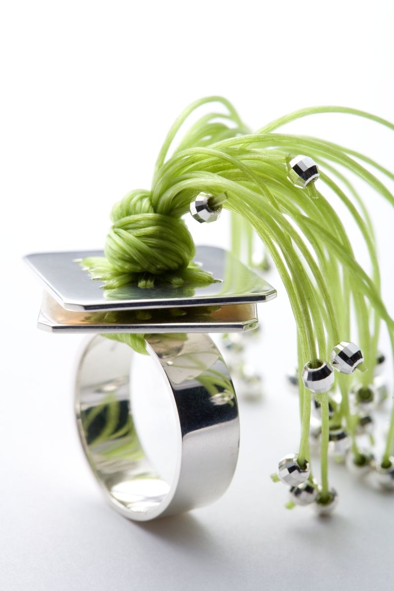 Medusa Ring. Material: Silber, Gummischnüre grün. Silberring mit zwei quadratischen Plättchen, durch die viele braune Gummischnüre gezogen sind und die in einem tentakelartigen Schweif auslaufen.