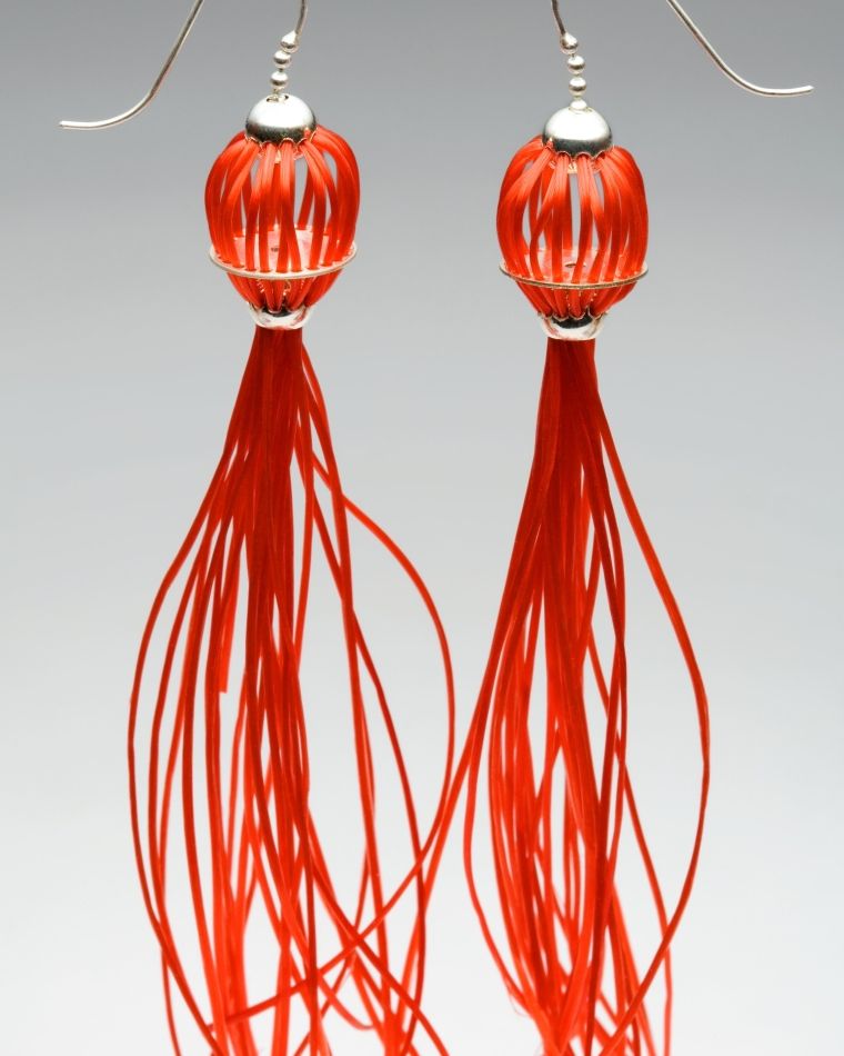 Medusa Ohrringe. Material: Silber, Gummischnüre rot. Der Körper dieser leichten Ohrringe wird durch viele rundlich angeordnete Gummischnüre geformt, die in einem tentakelartigen Schweif ausläuft.